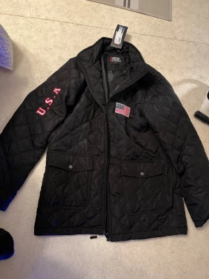 Svart quiltad jacka från Rogue - Quiltad svart jacka från Rogue Original med broderad USA-flagga på bröstet och texten 'U.S.A.' på ärmen. Jackan har två stora fickor framtill med knappar och dragkedja hela vägen. Perfekt för dig som vill ha en clean och sportig look.
