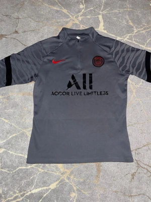 Grå PSG x Nike ströja - Säljer en grå Paris Saint-Germain träningströja från Nike med halv dragkedja. Tröjan har svarta och mörkgrå mönstrade ärmar, röd Nike-logga och PSG-märke på bröstet. Materialet är syntet och perfekt för fotbollsträning. Texten 'Accor Live Limitless' och 'Visit Rwanda' finns tryckt på framsidan och baksidan.