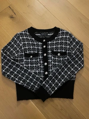 Svartvit rutig kofta från SHEIN - Säljer en svartvit rutig kofta från SHEIN med dekorativa silverknappar framtill och på fickorna. Koftan har lång ärm, rund halsringning och är stickad i ett mjukt material. Perfekt för dig som gillar klassisk men trendig stil.