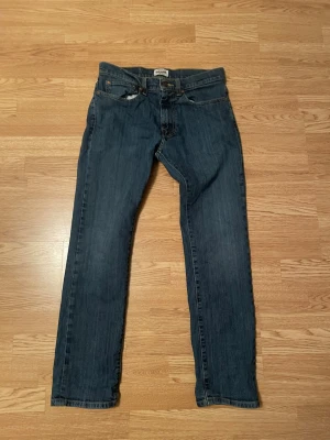 Blå raka jeans från Grant - Säljer ett par klassiska blå jeans från Grant i rak modell. Jeansen har fem fickor, orange kontrastsömmar och normal passform. Perfekta till vardagslooken och funkar året runt. Materialet är tåligt jeans-tyg i bomull. Små tecken på användning annars väldigt bra skick, sitter mer åt grisch hållet 