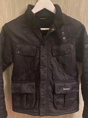 Svart quiltad jacka från Barbour - Säljer en svart quiltad jacka från Barbour med fyra fickor framtill, tryckknappar och dragkedja. Jackan har hög krage och Barbour-logga på ena ärmen. Klassisk och stilren design med rutmönstrad quiltning och detaljer i metall.
