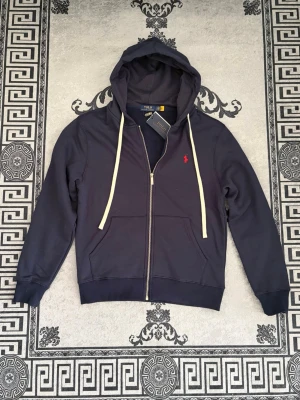 Polo kofta  - En marinblå zip-hoodie i pikématerial som kombinerar sportig elegans med vardaglig komfort. Tröjan har hel dragkedja, huva med dragsko och en stilren passform som passar perfekt för lager-på-lager.