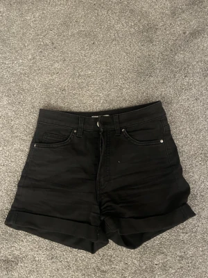Svarta high waist shorts från H&M - Säljer ett par svarta högmidjade shorts från H&M i storlek 36. Klassisk femficksmodell med uppvikta benslut och dragkedja framtill. Perfekta till sommaren och lätta att matcha med olika stilar.