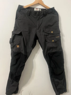Svarta cargopants från Fjällräven G-1000 - Säljer ett par svarta cargopants från Fjällräven i G-1000 material. Byxorna har flera stora fickor med tryckknappar, förstärkta knän och justerbara benslut. Perfekta för dig som gillar outdoor och vill ha slitstarka byxor med cool look.