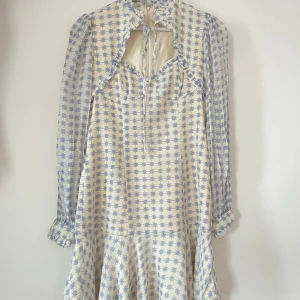 Blåvit blommig klänning By Malina - Supersöt kort klänning från By Malina i vitt med ljusblått blommönster. Klänningen har lång ärm, volangdetaljer och en snygg knytning vid halsen. Tillverkad i ett lätt och luftigt material, perfekt för varma dagar.