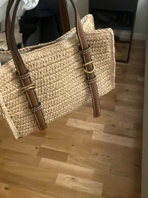 Beige axelväska i flätad strå från zara- slutsåld modell - Säljer en beige axelväska i flätad strå med bruna detaljer och handtag i konstläder. Väskan har guldfärgade metalldetaljer och en bred, avtagbar axelrem i beige tyg. Perfekt för sommarens alla äventyr!
