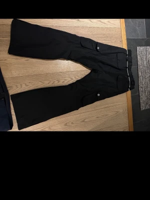 Svarta täckbyxor från Dope, storlek M - Säljer ett par svarta täckbyxor från Dope i storlek M. Byxorna har flera stora cargofickor med knappstängning och detaljer i vitt vid midjan. Tillverkade i slitstarkt syntetmaterial, perfekta för vinterns alla äventyr.