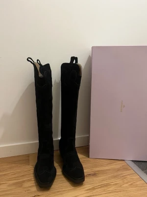Svarta mocka cowboy boots från Copenhagen - Säljer ett par höga svarta cowboy boots i mocka från Copenhagen. Bootsens design har klassiska broderade detaljer, fyrkantig tå och bred klack. De har dragflikar upptill för enkel påtagning och en riktigt snygg western-vibe. Oanvända 