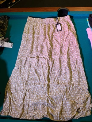 Beige blommig långkjol i bomull - Supersöt beige långkjol med vita och svarta blommor. Kjolen har resår i midjan för skön passform och är gjord i ett lätt bomullstyg som känns luftigt. Perfekt för dig som gillar en avslappnad men ändå trendig stil.