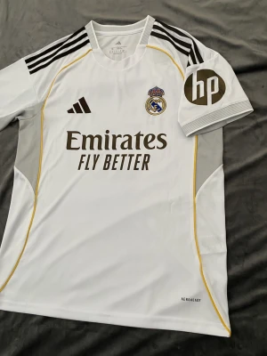 Real Madrid Bellingham matchtröja - Säljer en officiell Real Madrid matchtröja från Adidas med Bellingham och nummer 5 på ryggen. Tröjan är vit med svarta och gula detaljer, broderat klubbmärke och Adidas-logga, samt sponsortryck. Tillverkad i lätt och ventilerande polyester.