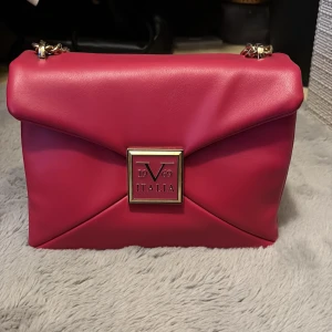 Rosa/röd axelväska från V1969 Italia - Snygg röd axelväska från V1969 Italia med guldfärgad kedjerem och dekorativ logga framtill. Väskan är tillverkad i slätt skinn och har flera fack inuti.                                                                            Väskan är ett dyrt märke, köptes för 1399kr på hemsida + frakten, tyvärr har den inte blivit använd och bara har varit i min garderob så nu hopas jag att den kan bli såld av en med denna stil ❣️ 