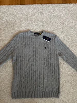 Grå kabelstickad tröja Polo Ralph Lauren - Säljer en grå kabelstickad tröja från Polo Ralph Lauren i storlek L. Tröjan har rund hals, långa ärmar och den klassiska broderade loggan på bröstet. Tillverkad i mjuk bomull och perfekt för lager på lager under höst och vinter. Aldrig använd! 