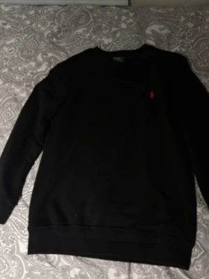 Svart sweatshirt från Polo Ralph Lauren - Klassisk svart sweatshirt från Polo Ralph Lauren i bomull med rund hals och diskret röd logga broderad på bröstet. Tröjan har långa ärmar och ribbade muddar vid ärmslut och nederkant. Perfekt för en clean och stilren look.