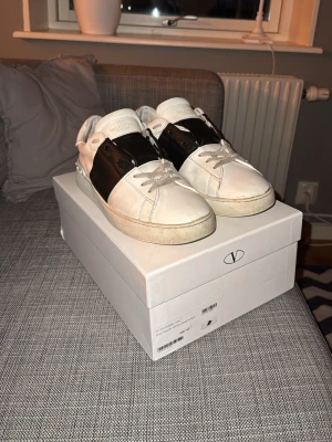 Valentino Garavani Open sneakers svart/vit - Säljer ett par Valentino Garavani Open sneakers i vit och svart skinn. Skorna har en bred svart läderpanel över mitten, vita snören och platt sula med rutmönster undertill. Klassisk logga på plösen och boxen medföljer. Riktigt snygga och stilrena sneakers. Skorna är i bra skick i säg men sätter ändå acceptabelt skick för vill inte få för mycket förväntningar 