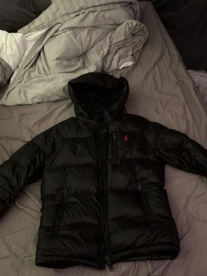 Svart pufferjacka från Polo Ralph Lauren - Säljer en svart pufferjacka från Polo Ralph Lauren med huva och dragkedja. Jackan har det klassiska röda Polo-logot på bröstet, tre fickor med dragkedja och justerbara muddar. Materialet är glansigt och känns riktigt modernt och fräscht. Pris kan diskuteras vid intresse