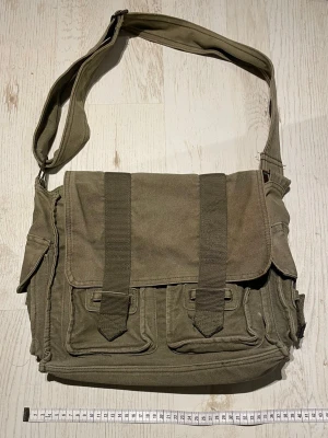 Axelväska olivgrön  - Rymlig olivgrön axelväska i slitstarkt material med flera ytterfickor och bred axelrem. Väskan har flera fickor framtill och på sidorna. Flertal missfärgningar se bild 3 & 4.