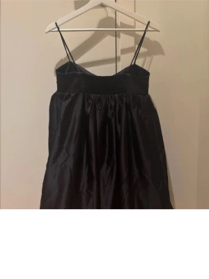 Svart kort klänning med tunna band - Säljer en svart kort klänning med tunna axelband och ballongliknande kjol. Klänningen har en markerad midja och lätt glansigt tyg som ger en elegant känsla. Perfekt för dig som vill ha en enkel men snygg look.