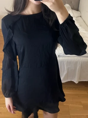 Svart volangklänning från divided H&M - Supersnygg svart kortklänning från Gina Tricot med volangdetaljer längs ärmar och nederkant. Klänningen har långa ärmar med resår vid handlederna och en rund halsringning. Storlek 36.