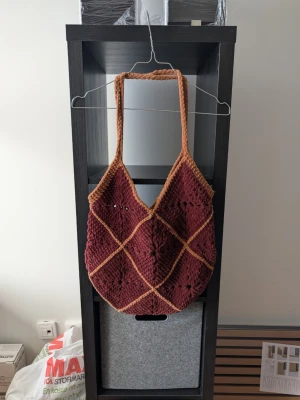 Handgjord brun stickad axelväska - Unik axelväska i vinröd och brun stickad design med rutmönster. Väskan har breda axelband och är gjord i mjukt garn, perfekt för dig som gillar en personlig och kreativ stil. Rymlig insida och snygga detaljer gör den till ett statement i vardagen.