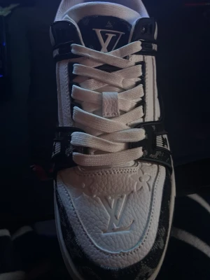 Louis Vuitton vita och svarta sneakers - Säljer ett par riktigt snygga Louis Vuitton sneakers i vitt och svart skinn med klassiskt LV-monogram och detaljer i svart. Skorna har snörning, platt sula och rund tå. Perfekta för dig som vill sticka ut med exklusiv streetstyle. Fick den i present men Den säljs för den passa inte mig för vi ville it förstöra den så den är bara använd en gång 