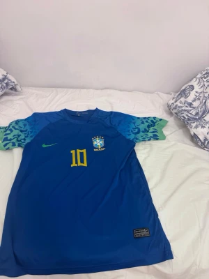 Blå Neymar Jr. Brasilien tröja - Säljer en blå Brasilien fotbollströja från Nike med Neymar Jr. och nummer 10 på ryggen. Tröjan har gula detaljer, grönt och blått mönster på ärmarna och Brasil-logga på bröstet. Perfekt för dig som älskar fotboll och vill stötta Brasilien.