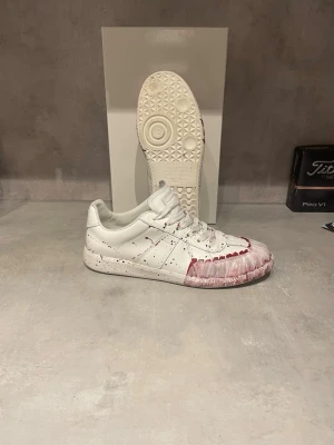 Maison Margiela - Unika vita sneakers i skinn med röda och svarta färgstänk och penseldrag över tåpartiet. Lågt skaft, snörning och klassisk siluett. Snygg detalj med färg på sulan och ovandelen. Perfekt för dig som vill sticka ut med ett kreativt uttryck.
