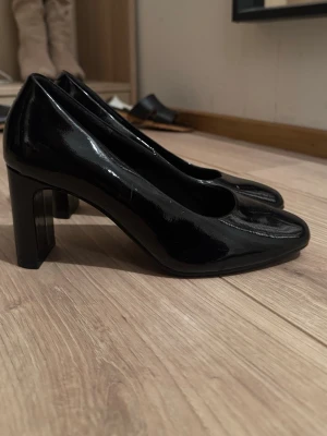 Svarta pumps från XIT med klack - Säljer ett par klassiska svarta pumps från XIT i glansigt material. Skorna har rund tå och stabil hög klack, perfekta för att lyfta vilken outfit som helst. Tillverkade i syntetmaterial med en stilren och tidlös look.