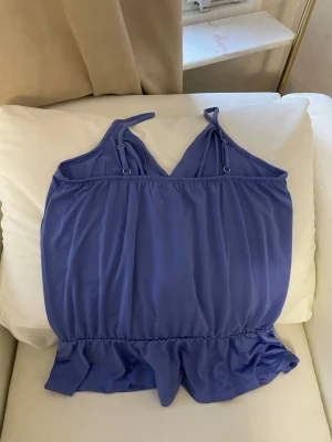 Ljuslila omlottopp från Ellos - Supersöt ljuslila omlottopp från Ellos med smala, justerbara axelband och volangkant nedtill. Toppen har en luftig passform och är tillverkad i ett mjukt, syntetiskt material som känns skönt mot huden. Perfekt för varma dagar och somriga vibes.