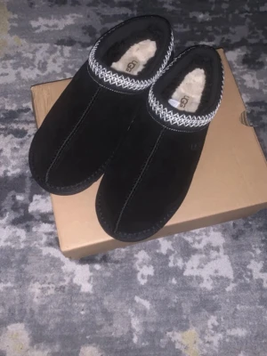 Svarta UGG mockatofflor med fårfoder - Mysiga svarta tofflor från UGG i mocka med fluffigt fårfoder och dekorativt mönster runt öppningen. Perfekta för kalla dagar hemma. Sulan är i slitstark gummi och insidan är supermjuk. Klassisk UGG-logga på innersulan.