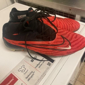 Nike Phantom GT2 röda fotbollsskor - Nike Phantom GT2 fotbollsskor i rött och svart med snyggt mönster och stora vita Swoosh-loggor på sidan och hälen. Skorna har snörning och en låg profil, perfekt för dig som vill ha grym touch och kontroll på planen. Ovandelen är i syntetmaterial och sulan har dobbar för gräs.
