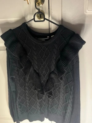 Svart stickad tröja med volang - Snygg svart stickad tröja med grovt kabelstickat mönster och breda volanger längs axlarna. Rund halsringning och långärmad modell som ger en cool och trendig vibe. Perfekt att styla med jeans eller kjol för en chill look.