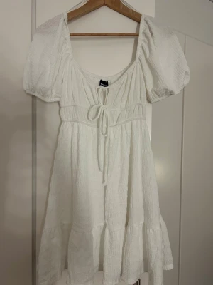 Vit kort klänning från Gina Tricot - Supersöt vit kort klänning från Gina Tricot i storlek XS. Klänningen har puffärmar, knytning framtill. Perfekt för sommaren. Använd 1 gång!