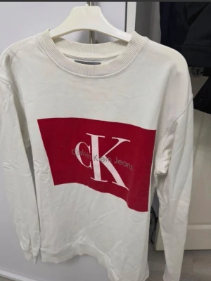 Vit sweatshirt Calvin Klein Jeans storlek s  - Vit sweatshirt från Calvin Klein Jeans med stor röd logga och CK-tryck framtill. Tröjan har rund halsringning, ribbade muddar och är långärmad. Regular fit och tillverkad i mjuk bomull för skön känsla. Stl S. En fläck.  se bild 5 och likt ett hål vid bild 6. Inget större märkbart därav pris.