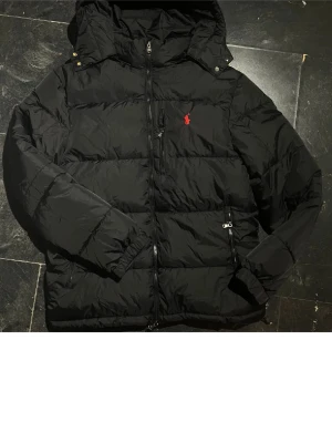 Svart pufferjacka från Polo Ralph Lauren - Säljer en svart pufferjacka från Polo Ralph Lauren med röd broderad logga på bröstet. Jackan har huva, flera dragkedjeförsedda fickor och justerbara detaljer vid ärmslut. Perfekt för kalla dagar och riktigt snygg till streetwear-stilen.