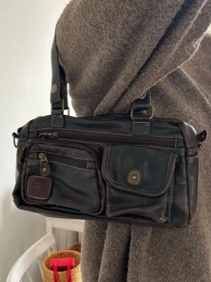 Mörkbrun axelväska i skinn - Säljer en mörkbrun axelväska i skinn med flera praktiska fack och detaljerade sömmar. Väskan har två rejäla axelband med nitar och spännen, samt dragkedja upptill och på framsidan. Perfekt för dig som vill ha en cool och funktionell väska med snygga detaljer.