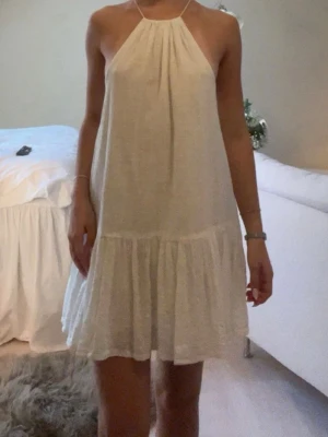 Vit kort sommarklänning med knyt - Luftig vit kortklänning med halterneck och knyt i nacken. Klänningen har en lös passform, volangkant nedtill och öppen rygg. 