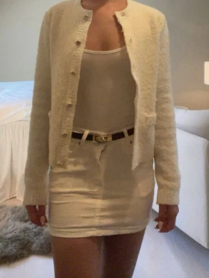 Off white kofta med guldknappar - Säljer en vit kofta i mjukt, strukturerat material med rund hals och dekorativa guldknappar framtill. Koftan har två små fickor fram och lång ärm. Passar även stl S