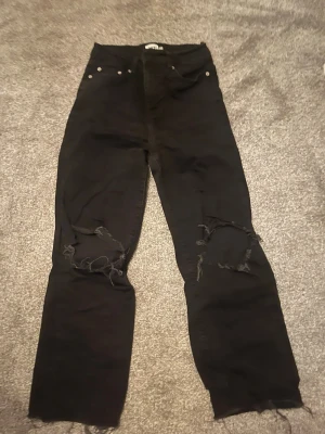 Svarta ripped jeans från lager 157 - Säljer ett par svarta jeans från lager 157 med höga knän och slitna detaljer över båda knäna. Byxorna har en rak passform och råa kanter vid bensluten för en extra edgy look. Perfekta för dig som gillar streetstyle och vill ha något unikt i garderoben.