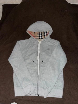 Burberry zipper - Burberry-hoodie med dragkedja. Sofistikerat grå nyans, accentuerad av det ikoniska Burberry-mönstret på huvan och längs insidan av dragkedjan. Material okänt, förmodligen en mjuk bomullsblandning. Storlek Medium. Full dragkedja fram, ribbstickade detaljer för en perfekt passform. En tidlös klassiker för en avslappnad lyxkänsla.