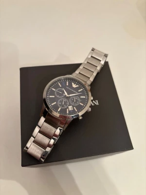 Emporio Armani klocka i stål - Snygg Emporio Armani klocka med silverfärgad länk i rostfritt stål och svart urtavla. Klockan har tre mindre urtavlor, datumvisning och klassisk logga upptill. Denna är oanvänd och i perfekt skick. Priset går att diskutera!