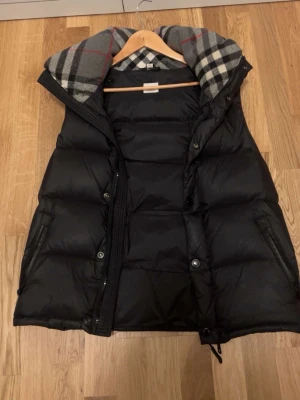 Svart Burberry pufferjacka med rutigt foder, bra skick  - Säljer en svart burberry pufferjacka med klassiskt rutigt foder i huvan. Jackan har dragkedja, knappar och två fickor med dragkedja. Perfekt för kyliga dagar och har en riktigt mysig vibe med sitt tjocka material och snygga detaljer. Bra skick! Kan både vara en väst och en pufferjacka.