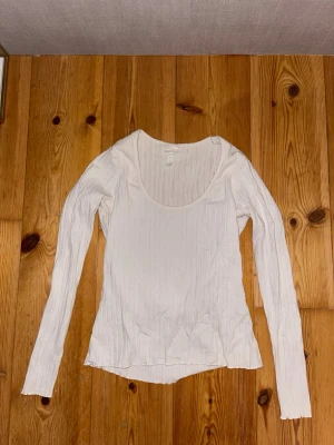 Vit ribbad långärmad topp H&M - Vit långärmad topp från H&M i storlek S. Toppen har ribbat mönster och en rundad, djup urringning. Tillverkad i mjukt material som känns skönt mot huden. Perfekt att styla med jeans eller kjol för en enkel och clean look.