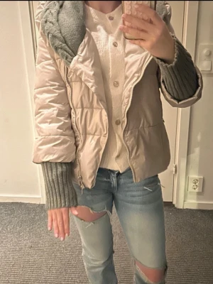 Beige pufferjacka med stickade detaljer - Säljer en beige pufferjacka med grå stickade muddar och krage. Jackan har dragkedja framtill och en mysig, quiltad look. Perfekt för kyliga dagar och ger en cool vibe till din outfit.