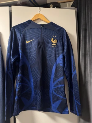 Nike Frankrike träningsset blå/guld - Säljer ett fett träningsset från Nike och Frankrikes landslag. Setet består av en långärmad träningströja med halv dragkedja och matchande byxor, båda i marinblått med guldiga detaljer och FFF-logga. Materialet är syntet och andas bra, perfekt för fotbollsträning.