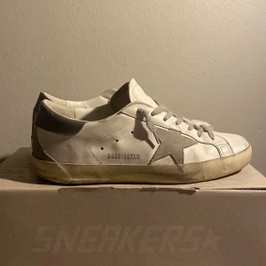Golden Goose Superstar sneakers vita/grå - Golden Goose Superstar sneakers i vitt skinn med grå mockadetaljer och stjärna på sidan. Skorna har grå snörning, beige sula och klassisk låg profil. Märkets logga syns på sidan och plösen. Perfekta för en cool och avslappnad stil. Pris kan diskuteras.
