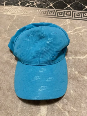 Blå Nike keps i polyester - Säljer en blå keps från Nike med justerbart spänne bak. Kepsen har all-over printat Nike-logga och är tillverkad i 100% polyester. Perfekt för dig som vill ha en sportig och clean look.