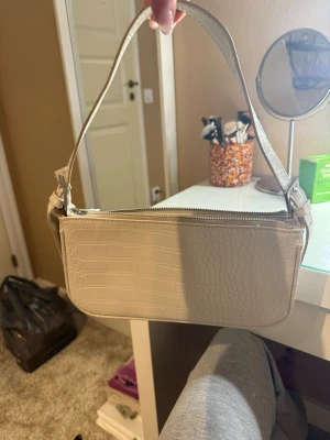 Beige axelväska från Gina Tricot - Snygg beige axelväska från Gina Tricot med croco-mönstrad yta och dragkedja upptill. Väskan har en stilren, rektangulär form och bred axelrem. Perfekt för att lyfta vilken outfit som helst med en trendig touch.