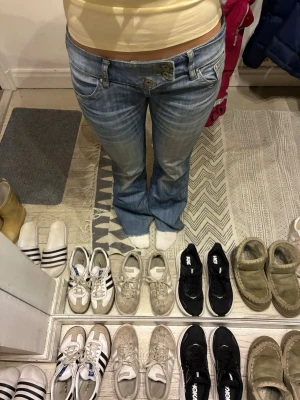 Lågmidjade bootcut jeans ljusblå - Säljer ett par riktigt snygga lågmidjade jeans i ljusblå tvätt med bootcut passform. Jeansen har slitna detaljer och dubbla knappar framtill för extra edge. Klassisk femficksmodell och tillverkade i jeansmaterial. De är köpta second hand i Danmark och märket är oklart