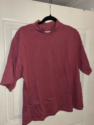 Vinröd t-shirt med hög krage Lindex - Enkel och stilren vinröd/smutsrosa t-shirt från Lindex med hög krage och avslappnad passform. Tillverkad i mjuk bomull och har korta ärmar. Perfekt för dig som gillar minimalistisk stil och vill ha något bekvämt till vardags.