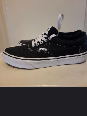 Svarta Vans sneakers med vit sula - Säljer ett par klassiska Vans sneakers i svart med vit sula och vita snören. Skorna har låg profil, rund tå och den ikoniska röda Vans-loggan på hälen. Ovandelen är i slitstark canvas och sulan har det typiska rutmönstret för extra grepp. Förvarats i förråd. Se bilder för skick. 